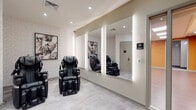 This single-floor wellness and fitness facility at 1000 Schindler Dr in South Amboy, NJ, is thoughtfully designed to offer a comprehensive health and relaxation experience. The space is divided into several distinct zones, each purposefully appointed for its function.

The massage and relaxation lounge is a standout area, featuring multiple full-body massage chairs set against textured, light-toned walls adorned with botanical artwork. A large, backlit mirror wall amplifies the sense of space and adds a sleek, spa-like ambiance. Light-toned stone-look tile flooring runs throughout this area, reinforcing the calming aesthetic.

Adjacent to the lounge is a bright, airy yoga and dance studio, distinguished by its vibrant orange accent walls and warm wood-look flooring. Wall-mounted ballet barres provide support for stretching and barre-style workouts, and panel skylights bathe the room in soft, diffused light.

The fitness room is well-equipped with cardio machines, including stationary bikes, alongside a full rack of exercise balls, resistance tools, and training accessories. Rubber flooring and bright recessed panel lighting create a functional, energizing environment.

Connecting these areas is a clean, neutral-toned corridor with stone-look tile flooring, built-in bench seating, and restroom access, ensuring smooth circulation throughout the facility. The overall design balances modern aesthetics with practical wellness functionality.