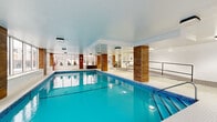 Located at 5501 Av Adalbert in Côte-Saint-Luc, Québec, this indoor aquatic facility offers a generously sized, single-level space designed for both recreational and lap swimming. The centerpiece is a large, luminous pool with vivid turquoise water, framed by mosaic tile edging and equipped with underwater lighting and stainless steel entry rails for safe access. A separate hot tub area is also present, providing a dedicated space for relaxation.

The facility's design balances function with aesthetic appeal. Warm wood-clad columns punctuate the space at regular intervals, providing a striking contrast against the crisp white walls and smooth ceiling. A continuous band of large, floor-to-ceiling windows runs along multiple walls, flooding the interior with natural light and offering views of the surrounding outdoor environment. The flooring throughout the pool surround features classic hexagonal white mosaic tiles, lending a clean and timeless look.

A raised entry area with metal railings and steps provides a transition point into the pool deck, with posted safety regulations clearly visible. The space also includes a dedicated entry with sliding glass doors. Overall, the facility presents a well-maintained, modern aquatic environment suited for year-round use.