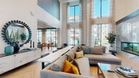 This sophisticated single-floor amenity space is designed with a strong emphasis on modern aesthetics and community living. The centerpiece of the space is a dramatic double-height lounge area bathed in natural light streaming through expansive floor-to-ceiling windows adorned with sheer linen drapes. The lounge features herringbone-patterned wood-look tile flooring, a sleek built-in fireplace framed in polished stone, and a wall-mounted television set within a custom blue-accented built-in shelving unit — creating a refined yet welcoming atmosphere. Comfortable sectional sofas with mustard and patterned throw pillows invite residents to relax and socialize. A social kitchen and bar area showcases bold slate-blue walls, a large quartz-topped island with bar seating, industrial-style pendant lighting, and dual wall-mounted televisions, making it ideal for gatherings and events. Glass-panel sliding doors separate distinct zones within the space while maintaining visual openness and connectivity. Intimate seating nooks positioned beside floor-to-ceiling windows offer views of lush landscaped courtyards. A private conference room, enclosed by glass partitions, features a live-edge wood table, sculptural pendant chandelier, and wall-mounted display — perfectly suited for professional or private meetings. Throughout, the design palette of soft blues, warm wood tones, and crisp whites creates a cohesive and elevated ambiance.