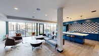 This single-floor space combines a fully equipped kitchen and an open living area in a thoughtfully designed layout. The kitchen is a standout feature, showcasing bold navy-blue cabinetry paired with a striking geometric patterned tile backsplash. A long white-quartz island with bar-height seating serves as the kitchen's centerpiece, illuminated by sleek pendant lights hanging overhead. Professional-grade appliances, including a multi-burner gas range, reinforce the kitchen's functional yet stylish character.

The living area flows seamlessly from the kitchen, offering generous space for seating arrangements anchored by a sectional sofa and a sculptural round coffee table atop a woven area rug. Open shelving along one wall provides display and storage, while a wall-mounted television adds a media-focused element to the lounge area.

Throughout the space, light wood-look flooring creates a warm, cohesive foundation. Floor-to-ceiling windows and glass sliding doors line one wall, flooding the interior with abundant natural light and providing direct views of and access to an outdoor terrace. Accent walls in soft teal add color and depth, complemented by framed artwork. Recessed ceiling lighting ensures the space is well-lit throughout.