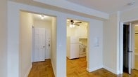 This single-floor, one-bedroom, one-bathroom residence presents a clean and functional layout with notable classic details throughout. The entryway opens to a hallway with warm parquet wood flooring laid in a traditional herringbone pattern, which extends into the adjoining spaces and adds a timeless character to the interior.

The kitchen is accessed through a wide arched opening framed by structural columns, giving the space a defined yet open feel. It is equipped with white cabinetry, a white refrigerator, and a white gas range, all set beneath a ceiling fan that provides ventilation and ambient lighting. The cabinetry extends to the upper walls, offering ample storage. A small arched niche on the wall adds an architectural accent detail reminiscent of pre-war design sensibilities.

The overall color palette is light and neutral, with white walls and trim throughout, contributing to a bright and airy atmosphere. The space is well-proportioned for a one-bedroom unit, with clearly delineated zones for entry, cooking, and living. The combination of parquet flooring and arched architectural detailing gives the residence a classic urban character.