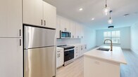 Located at 5025 Huntly Ridge St in Charlottesville, VA, this well-appointed one-bedroom, one-bathroom residence offers a thoughtfully designed layout with modern finishes throughout. The open-concept kitchen and living area serves as the heart of the home, featuring crisp white shaker cabinetry with matte black hardware, stainless steel appliances including a full-size refrigerator, range, and microwave, and a clean white countertop island with a pendant light fixture. A white subway tile backsplash and light wood-style flooring complete the contemporary aesthetic of the kitchen. The generously sized bedroom is carpeted for comfort, fitted with a ceiling fan, and features a large window that brings in natural light and views of the surrounding neighborhood. A dedicated walk-in closet with wire shelving provides ample storage directly accessible from the bedroom. The full bathroom is finished in the same clean, modern palette, offering a vanity with white cabinetry and black hardware, a tub/shower combo, and light wood flooring that carries through from the main living areas. Throughout the residence, neutral tones, recessed lighting, and consistent design details create a cohesive and move-in-ready living environment.