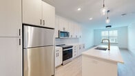Located at 5025 Huntly Ridge St in Charlottesville, VA, this well-appointed one-bedroom, one-bathroom residence offers a thoughtfully designed layout with modern finishes throughout. The open-concept kitchen and living area serves as the heart of the home, featuring crisp white shaker cabinetry with matte black hardware, stainless steel appliances including a full-size refrigerator, range, and microwave, and a clean white countertop island with a pendant light fixture. A white subway tile backsplash and light wood-style flooring complete the contemporary aesthetic of the kitchen. The generously sized bedroom is carpeted for comfort, fitted with a ceiling fan, and features a large window that brings in natural light and views of the surrounding neighborhood. A dedicated walk-in closet with wire shelving provides ample storage directly accessible from the bedroom. The full bathroom is finished in the same clean, modern palette, offering a vanity with white cabinetry and black hardware, a tub/shower combo, and light wood flooring that carries through from the main living areas. Throughout the residence, neutral tones, recessed lighting, and consistent design details create a cohesive and move-in-ready living environment.