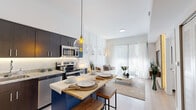 This model unit at 2333 NW 167th St in Miami Gardens presents a thoughtfully designed two-bedroom, one-bathroom layout that maximizes every inch of its compact single-floor footprint. The open-concept kitchen and living area form the heart of the unit, where soaring ceilings amplify the sense of space. The kitchen is equipped with stainless steel appliances, dark espresso cabinetry with under-cabinet lighting, granite countertops, a subway tile backsplash, and a navy blue island with a butcher block top that doubles as a casual dining space beneath a globe pendant light.

The living room flows seamlessly from the kitchen, featuring a textured stone accent wall with a mounted entertainment unit, white wainscoting panels, and floor-to-ceiling sliding glass doors that flood the space with natural light. Both bedrooms are bright and serene, with sheer curtains diffusing sunlight and light wood-tone flooring underfoot. The primary bedroom features a decorative accent wall, while the second bedroom offers a clean, minimalist design.

The bathroom includes a granite-topped vanity with dark cabinetry, a tub-shower combination, and light wood-tone flooring, maintaining the unit's cohesive, contemporary aesthetic throughout.