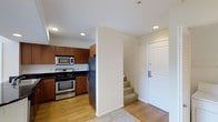 This two-story residence offers a well-organized layout that efficiently connects its living spaces across both floors. The entry level is immediately welcoming, featuring a clean, minimalist foyer with light hardwood flooring and neutral cream-toned walls that set a bright, airy tone throughout.

The kitchen is a standout feature on this level, outfitted with rich warm-toned wood cabinetry that extends to the upper walls, providing generous storage capacity. Dark countertops offer a strong visual contrast against the cabinetry, while a full suite of stainless steel appliances — including a gas range with oven, built-in microwave, and a full-sized refrigerator — equips the space for practical, everyday use. Recessed lighting overhead ensures the kitchen remains well-lit and functional.

Adjacent to the kitchen, the entryway provides a seamless transition into the space, with a solid white entry door and a staircase leading directly to the upper floor. The stairs are carpeted, offering a subtle distinction in material from the hardwood flooring below. The overall ambiance is clean, modern, and practical, with a neutral palette that allows for versatile interior styling across both floors.