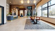 Located at 1201 W Grove St in Boise, Idaho, this sophisticated lobby spans a generously sized single floor, offering a seamless blend of modern luxury and welcoming warmth. The grand entrance features a striking floor-to-ceiling glass façade framed in dark metal, flooding the interior with natural light and offering views of the surrounding streetscape. Large-format stone tile flooring flows throughout, unifying the space with a clean, refined aesthetic.

At the heart of the lobby sits a circular seating island with channel-tufted navy upholstery, topped by an artistic branching chandelier that serves as a dramatic focal point. Soaring ceilings with warm wood-toned panels add architectural depth, while an array of designer pendant and globe fixtures layer the space with ambient and accent lighting.

The front desk, branded "Saratoga," anchors one side of the lobby alongside a communal work table with bold red seating. A lounge area features a floor-to-ceiling marble fireplace flanked by cognac leather chairs, alongside a curated bar display. Banquette seating with large windows provides a casual dining nook, while arched passageways lead to additional lounge zones with eclectic, colorful furnishings. Lush indoor greenery is thoughtfully integrated throughout, enhancing the inviting, upscale ambiance.