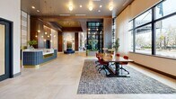 Located at 1201 W Grove St in Boise, Idaho, this sophisticated lobby spans a generously sized single floor, offering a seamless blend of modern luxury and welcoming warmth. The grand entrance features a striking floor-to-ceiling glass façade framed in dark metal, flooding the interior with natural light and offering views of the surrounding streetscape. Large-format stone tile flooring flows throughout, unifying the space with a clean, refined aesthetic.

At the heart of the lobby sits a circular seating island with channel-tufted navy upholstery, topped by an artistic branching chandelier that serves as a dramatic focal point. Soaring ceilings with warm wood-toned panels add architectural depth, while an array of designer pendant and globe fixtures layer the space with ambient and accent lighting.

The front desk, branded "Saratoga," anchors one side of the lobby alongside a communal work table with bold red seating. A lounge area features a floor-to-ceiling marble fireplace flanked by cognac leather chairs, alongside a curated bar display. Banquette seating with large windows provides a casual dining nook, while arched passageways lead to additional lounge zones with eclectic, colorful furnishings. Lush indoor greenery is thoughtfully integrated throughout, enhancing the inviting, upscale ambiance.
