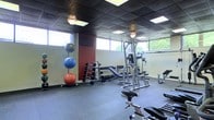 This single-floor fitness center offers a practical and functional workout environment designed to accommodate a variety of exercise routines. The space is outfitted with rubber flooring throughout, providing both safety and durability underfoot. A dark drop ceiling with recessed panel lighting keeps the area well-lit, while a row of windows along the upper walls allows natural light to filter in, creating an open and airy atmosphere.

The strength training section is well-appointed, featuring a multi-station cable and weight machine suitable for a wide range of resistance exercises. A full dumbbell rack with a graduated selection of free weights sits alongside an adjustable workout bench, offering versatility for targeted strength training. A vertical rack of stability and medicine balls in varying sizes adds functional training options to the space.

For cardio, stationary bikes are positioned toward the front of the room, providing low-impact aerobic workout options. The combination of machines, free weights, and functional fitness equipment makes this a comprehensive facility. The overall ambiance is no-frills and purpose-driven, with every element oriented toward maximizing the usability of the space for fitness enthusiasts of all levels.