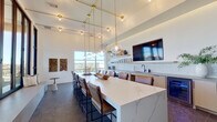 This elegantly designed lounge is a single open-plan space that masterfully blends modern aesthetics with functional versatility. Soaring ceilings amplify the sense of volume, while a dramatic grid of floor-to-ceiling black-framed glass walls floods the interior with abundant natural light and frames sweeping panoramic views of the surrounding cityscape and landscape.

At the heart of the space sits a generous white quartz island with bold veining, surrounded by leather bar-height chairs — a natural focal point for gathering and socializing. Overhead, a sculptural brass pendant lighting system suspends globe lights at varying heights, adding warmth and a refined industrial character to the room.

Along one wall, a well-appointed service area features open floating shelves stocked with dishware, a white tile backsplash, and light wood cabinetry with a clean, minimalist finish. A mounted flat-screen TV and a matte-finish countertop complete this functional zone.

A built-in cushioned bench seat nestled against the glass wall offers a cozy nook with direct access to an outdoor terrace. A bold navy blue accent wall with vertical shiplap paneling and curated wall art anchors the opposite end of the room, adding depth and contrast. Dark large-format tile flooring grounds the entire space with a sleek, polished finish.