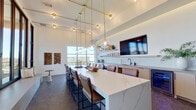 This elegantly designed lounge is a single open-plan space that masterfully blends modern aesthetics with functional versatility. Soaring ceilings amplify the sense of volume, while a dramatic grid of floor-to-ceiling black-framed glass walls floods the interior with abundant natural light and frames sweeping panoramic views of the surrounding cityscape and landscape.

At the heart of the space sits a generous white quartz island with bold veining, surrounded by leather bar-height chairs — a natural focal point for gathering and socializing. Overhead, a sculptural brass pendant lighting system suspends globe lights at varying heights, adding warmth and a refined industrial character to the room.

Along one wall, a well-appointed service area features open floating shelves stocked with dishware, a white tile backsplash, and light wood cabinetry with a clean, minimalist finish. A mounted flat-screen TV and a matte-finish countertop complete this functional zone.

A built-in cushioned bench seat nestled against the glass wall offers a cozy nook with direct access to an outdoor terrace. A bold navy blue accent wall with vertical shiplap paneling and curated wall art anchors the opposite end of the room, adding depth and contrast. Dark large-format tile flooring grounds the entire space with a sleek, polished finish.