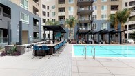 This expansive outdoor amenity courtyard is designed to deliver a resort-caliber experience. At its center sits a generously sized swimming pool with crystal-clear blue water, flanked by wide concrete pool decking that provides ample space for movement and relaxation. A separate spa is tucked along the perimeter, enclosed by sleek teal privacy screens that add a modern, architectural accent to the space.

Anchoring one side of the courtyard is a covered outdoor bar area, featuring a long counter-height surface lined with contemporary blue wire-frame barstools. A dark steel pergola overhead provides shade while maintaining an open-air ambiance. Adjacent to the bar, additional pergola-shaded seating zones offer cushioned lounge chairs and casual furniture, ideal for social gatherings or quiet relaxation.

Tall palm trees rise throughout the courtyard, lending a warm, Southern California aesthetic and framing the multi-story residential building that wraps around the space. The building's balconies overlook the courtyard, creating a dynamic sense of enclosure without sacrificing openness.

Paved walkways with a patterned brick-style finish connect all areas of the courtyard, complemented by native landscaping and ornamental plantings. An outdoor shower station with matte black fixtures and tile surround provides functional poolside convenience. The overall design blends modern urban architecture with leisure-focused outdoor living.