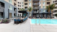 This expansive outdoor amenity courtyard is designed to deliver a resort-caliber experience. At its center sits a generously sized swimming pool with crystal-clear blue water, flanked by wide concrete pool decking that provides ample space for movement and relaxation. A separate spa is tucked along the perimeter, enclosed by sleek teal privacy screens that add a modern, architectural accent to the space.

Anchoring one side of the courtyard is a covered outdoor bar area, featuring a long counter-height surface lined with contemporary blue wire-frame barstools. A dark steel pergola overhead provides shade while maintaining an open-air ambiance. Adjacent to the bar, additional pergola-shaded seating zones offer cushioned lounge chairs and casual furniture, ideal for social gatherings or quiet relaxation.

Tall palm trees rise throughout the courtyard, lending a warm, Southern California aesthetic and framing the multi-story residential building that wraps around the space. The building's balconies overlook the courtyard, creating a dynamic sense of enclosure without sacrificing openness.

Paved walkways with a patterned brick-style finish connect all areas of the courtyard, complemented by native landscaping and ornamental plantings. An outdoor shower station with matte black fixtures and tile surround provides functional poolside convenience. The overall design blends modern urban architecture with leisure-focused outdoor living.