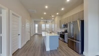 This single-story space is designed with a clean, modern aesthetic and an open, functional layout. The kitchen serves as the centerpiece of the main living area, featuring a large center island with an integrated sink and white quartz countertop, gray shaker-style cabinetry extending to the ceiling, and a full suite of stainless steel appliances including a gas range, built-in microwave, and French door refrigerator. Recessed lighting and light wood-look luxury vinyl plank flooring run throughout the open-concept main area, which flows toward a front entry door with windows allowing natural light to filter in. A ceiling fan is positioned in the living area, adding to the airy feel of the space.

The bedrooms are carpeted and offer a quiet, comfortable atmosphere, with windows fitted with blinds for light control. At least one bedroom includes a ceiling fan. A back door with a glass panel provides additional access and natural light to one of the secondary rooms.

The bathrooms are finished with striking marble-style tile covering both the floors and shower walls. Each bathroom features a gray vanity with white countertop, brushed nickel fixtures, and a large mirror. The primary bathroom includes a glass-enclosed walk-in shower, while a secondary bathroom offers a streamlined vanity and toilet configuration.