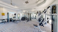 Located at 4132 53rd Ave W in Bradenton, Florida, this single-story amenity facility is thoughtfully designed to offer both active and leisure-focused spaces for its users. The layout divides seamlessly into two primary zones: a comprehensive fitness center and a modern recreation and lounge area.

The fitness center is spacious and well-lit, benefiting from abundant natural light streaming through multiple large windows and French doors that frame views of the surrounding greenery. A tray ceiling with a ceiling fan adds architectural interest overhead, while floor-to-ceiling mirrors visually expand the room. The space is outfitted with cardio equipment including treadmills and ellipticals, a cable machine, a full dumbbell rack, and weight benches, all set atop patterned, multi-tone carpeted flooring.

The adjacent recreation area transitions to light wood-look flooring and features a coastal-inspired aesthetic with sage green accent walls. It includes a foosball table, a bistro-style seating nook with large windows overlooking lush landscaping, and a cozy lounge corner with a wall-mounted TV, floating wood shelves, and upholstered seating. A wet bar equipped with a sink, beverage cooler, and dark cabinetry, alongside a wall of storage lockers, adds practical convenience to the space.