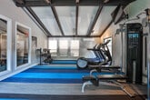 Located at 777 Valleybrook Crossing in Decatur, Georgia, this well-appointed fitness center offers a thoughtfully designed space for residents to pursue their wellness goals. The single-level layout is defined by striking vaulted ceilings with exposed dark wood beams that add architectural character and a sense of height and openness throughout the room.

The flooring features a bold, striped carpet in shades of blue, gray, and teal, creating a dynamic and energizing atmosphere. Large windows line multiple walls, flooding the space with abundant natural light and offering views of the surrounding greenery and community grounds, contributing to an inviting, open feel.

The facility is equipped with a comprehensive selection of fitness equipment, including treadmills, elliptical machines, and a cable machine system with an adjustable bench for strength training. A dedicated rack holds a variety of medicine balls and kettlebells, supporting functional fitness routines. Full-length mirrors are positioned along one wall, enhancing the sense of space while providing visual feedback during workouts. A wall-mounted flat-screen television adds entertainment for cardio sessions.

The walls are finished in a clean white shiplap style, complementing the exposed beam ceiling and contributing to the gym's modern yet warm aesthetic. The overall ambiance is bright, spacious, and well-equipped, making it a practical and pleasant environment for daily exercise.