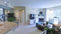 This single-floor space features an open-concept living area finished in warm neutral tones, with light beige walls and plush wall-to-wall carpeting that contribute to a comfortable, inviting atmosphere. The living room serves as the focal point of the space, centered around a clean white fireplace that adds a classic architectural detail to the room. A flat-screen television is mounted on a white media console positioned adjacent to the fireplace, creating a functional and cohesive entertainment area. Decorative touches, including potted greenery, a large wall clock, and blue accent vases displayed on the fireplace mantle, lend personality to the space. A wooden coffee table anchors the seating area, while natural light filters into the room through a window dressed with patterned curtains. The open layout connects the living area to adjacent spaces, including what appears to be a dining area with a granite-topped surface visible in the foreground. A hallway leading to additional rooms is visible in the background, with a white paneled door offering a sense of depth and flow throughout the layout. HVAC vents are integrated neatly into the walls and floors.