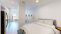 This single-floor residence showcases a thoughtfully designed open-concept layout that seamlessly connects its key living areas. The living room is a standout, featuring soaring ceilings with exposed white-painted beams that lend an airy, loft-like character to the space. Large windows invite abundant natural light and offer views of the surrounding urban streetscape, complementing the light hardwood flooring found throughout. The kitchen is defined by a striking two-tone design, pairing dark lower cabinetry with crisp white upper cabinets and a classic white square-tile backsplash. A full suite of appliances, including a refrigerator and microwave, provides full functionality within the compact, well-organized space. The bedroom is a calm, minimal retreat enclosed by clean white walls and accessed via a half-wall divider fitted with an open cube shelving unit that subtly separates it from the living area while maintaining an open flow. The room accommodates a full-sized bed and features a full-length mirror and patterned area rug for added warmth. The bathroom continues the minimalist aesthetic with all-white subway tile surrounding a full bathtub, accented by sleek matte black fixtures. The overall ambiance is modern, light-filled, and cohesive.
