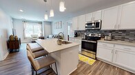 The Hickory floor plan in Ann Arbor, Michigan offers a thoughtfully designed single-level layout with two bedrooms and two full bathrooms. The open-concept kitchen and living area serves as the heart of the home, featuring wood-look flooring throughout. The kitchen is outfitted with white shaker-style cabinetry, a mosaic tile backsplash, stainless steel appliances including a range, microwave, and refrigerator, and a central island with an undermount sink and bar seating illuminated by pendant lighting. The living area flows seamlessly from the kitchen, creating a cohesive and functional shared space.

Both bedrooms are carpeted and receive natural light through large windows with exterior views. The rooms offer generous proportions with neutral walls that complement a variety of furnishing styles. The two full bathrooms feature wood-look tile flooring, dark espresso-toned vanities with white countertops, and clean, neutral walls. One bathroom includes a tiled shower-tub combination, while the other provides a more streamlined layout. The overall aesthetic throughout the unit is contemporary and cohesive, with a light, neutral color palette that creates a bright and welcoming atmosphere.