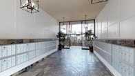 This lobby serves as an elegant and functional entry point to the building. The space is finished with a herringbone-patterned stone tile floor in cool grey and blue tones, lending a polished, upscale character to the entryway. On both sides of the corridor, built-in mailbox banks finished in light grey are framed by white wainscoting and stone veneer bases, providing a clean, organized appearance for residents. The white paneled walls rise to a warm-toned ceiling, creating a bright and airy atmosphere throughout the hall. Pendant lantern-style light fixtures hang at measured intervals, offering warm ambient lighting that complements the natural light streaming in from the front. Floor-to-ceiling glass doors and sidelights at the far end of the lobby provide an unobstructed view to the exterior courtyard, where steps, mature palm trees, and greenery are visible. Flanking the glass entry are potted green plants that introduce a natural, welcoming element to the space. Overall, the lobby balances practicality with an inviting aesthetic, making for a well-maintained and attractive communal entrance.