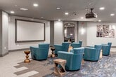 This screening room offers a dedicated home theater experience with a thoughtfully designed, modern aesthetic. The room is anchored by a large, framed projection screen mounted on a gray accent wall, complemented by a ceiling-mounted projector for high-quality viewing. Recessed lighting throughout the dark ceiling creates a cinematic ambiance while providing functional illumination when needed.

Seating consists of numerous teal upholstered swivel lounge chairs arranged in a loose, tiered configuration across the room, ensuring unobstructed sightlines to the screen. Decorative wooden side tables of varying styles are interspersed between the chairs, offering convenient surfaces for guests. The flooring features a combination of patterned carpet in the primary seating area and neutral-toned carpet tiles, adding visual interest and defining different zones within the space.

The rear of the room includes a casual bar-height table with stools, ideal for those who prefer a more upright seating option. Wall sconces along the navy blue accent wall at the back provide warm ambient lighting. An arched doorway leads to an adjacent lounge or common area, enhancing the overall flow and accessibility of the space.