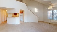 This two-floor residence showcases an open, airy design anchored by a dramatic vaulted ceiling in the main living area. The living room is generously proportioned, featuring plush carpeting, a classic fireplace, and a sliding glass door that opens to an outdoor area, flooding the space with natural light and garden views. Clerestory windows positioned high along the vaulted walls further enhance the bright, open ambiance. A staircase with a wood handrail leads to the upper level, visible from the main living space and overlooked by a loft-style landing. The kitchen is efficiently laid out with warm oak cabinetry, light countertops, tile flooring, and white appliances including a range, microwave, and refrigerator. The upper floor accommodates carpeted bedrooms, including one with angled ceilings that follow the roofline and louvered bifold closet doors providing ample storage. Additional bedrooms on the main level offer neutral carpeting and natural light through blinds-fitted windows. The bathroom is clean and functional, finished with tile flooring, a tub-shower combination, and a vanity with warm wood cabinetry. Throughout the home, a neutral color palette of whites and warm beiges creates a cohesive, move-in-ready atmosphere suited to a variety of interior styles.