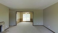 This single-floor space presents a practical and well-connected layout designed for comfortable everyday living. The interior is finished with neutral gray and taupe wall tones throughout, complemented by light-colored carpet flooring that adds warmth underfoot and ties the spaces together cohesively. Dark-toned trim and baseboards provide a subtle contrast, framing each room with a grounded, traditional aesthetic. The main living area flows openly into an adjoining room through a wide, defined entryway opening, creating a sense of connectivity between spaces while still maintaining a sense of separation. The adjoining room features a ceiling fan, offering both practical air circulation and a classic finishing touch. A large window in this secondary room allows natural light to pour in, brightening the space and offering a view of the surrounding greenery outside. The overall ambiance is clean, understated, and move-in ready, with a neutral palette that provides an excellent canvas for personalization. The single-floor layout ensures easy accessibility and a straightforward flow from one area to the next, making the space both functional and inviting.