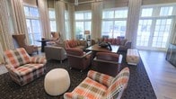 This sophisticated lounge space is designed to offer comfort and style in equal measure. The room is anchored by large windows dressed with floor-to-ceiling cream drapes that allow generous natural light to flood the interior, offering pleasant views of the surrounding greenery. The seating arrangement is thoughtfully curated, featuring a mix of plush leather sofas and armchairs in warm earth tones — including rich browns, deep rusts, and bold plaid patterns — that lend the space a cozy yet refined character. Accent pillows in terracotta and orange add vibrant pops of color throughout. A sturdy wooden coffee table serves as the centerpiece of the main seating cluster, complemented by a tufted ottoman and a round pouf that add textural variety. Subtle overhead lighting, including a chandelier visible near the ceiling, enhances the room's ambiance during evening hours. The patterned carpet grounds the space and ties the warm color palette together seamlessly. Overall, this lounge exudes an inviting, upscale atmosphere well-suited for relaxed conversation, casual gatherings, or quiet moments of leisure.