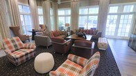 This sophisticated lounge space is designed to offer comfort and style in equal measure. The room is anchored by large windows dressed with floor-to-ceiling cream drapes that allow generous natural light to flood the interior, offering pleasant views of the surrounding greenery. The seating arrangement is thoughtfully curated, featuring a mix of plush leather sofas and armchairs in warm earth tones — including rich browns, deep rusts, and bold plaid patterns — that lend the space a cozy yet refined character. Accent pillows in terracotta and orange add vibrant pops of color throughout. A sturdy wooden coffee table serves as the centerpiece of the main seating cluster, complemented by a tufted ottoman and a round pouf that add textural variety. Subtle overhead lighting, including a chandelier visible near the ceiling, enhances the room's ambiance during evening hours. The patterned carpet grounds the space and ties the warm color palette together seamlessly. Overall, this lounge exudes an inviting, upscale atmosphere well-suited for relaxed conversation, casual gatherings, or quiet moments of leisure.