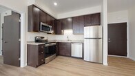 This single-floor unit is designed with a clean, modern aesthetic that emphasizes light and functionality throughout. The main living area is generously proportioned, featuring light-toned hardwood flooring that runs seamlessly throughout the entire unit. Natural light streams in through a multi-pane window overlooking lush greenery, creating a bright and airy ambiance. A ceiling fan is centrally mounted overhead, and a wall-mounted mini-split air conditioning unit provides efficient climate control. Recessed lighting and crisp white walls with white baseboards contribute to the space's fresh, contemporary feel.

The kitchen is well-equipped and thoughtfully designed, showcasing rich dark-brown shaker-style cabinetry that extends to the ceiling for ample storage. Light granite countertops provide a striking contrast, and the full suite of stainless steel appliances includes a refrigerator, range with oven, microwave, and dishwasher. The kitchen opens directly to the main living area, creating a cohesive, open-concept flow.

The bathroom continues the unit's consistent design language, featuring the same hardwood-style flooring, a dark-toned vanity with a white countertop, and a full glass-enclosed shower stall, offering both practicality and style.