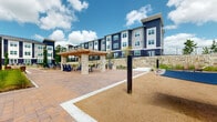Located at 3272 State Highway 30 in Huntsville, Texas, this expansive outdoor amenity courtyard is designed to provide residents with a vibrant and functional community gathering space. The courtyard is framed by a striking three-story building featuring a bold navy blue and white board-and-batten exterior with black iron railings and natural stone retaining walls, lending the space a modern yet inviting aesthetic.

At the heart of the courtyard, a handsome pergola supported by natural stone pillars and warm wood beams serves as a central outdoor dining and gathering area, complete with seating and a nearby grilling station. String lights overhead add ambiance for evening use. Adjacent to the pergola, a bocce ball court with artificial turf provides a dedicated recreational area, flanked by Adirondack-style seating.

A dedicated hammock garden, set within a gravel and landscaped zone, offers a relaxed retreat with multiple suspended hammocks. Brick-paver pathways connect the various activity zones, creating a cohesive and well-organized layout throughout the grounds. Ornamental grasses, young shade trees, and native plantings are thoughtfully integrated throughout, enhancing the natural character of the space. The open-air design maximizes sunlight while providing a welcoming outdoor environment for leisure and socializing.
