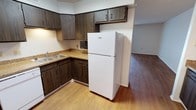 This single-floor space offers a practical and comfortable layout centered around a well-equipped kitchen. The kitchen features dark wood cabinetry on both the upper and lower levels, providing ample storage for everyday needs. A double-basin sink sits beneath the upper cabinets, complemented by earthy, speckled countertops that offer generous prep space. The kitchen is equipped with a full-size top-freezer refrigerator and includes space for additional appliances, with a compact microwave tucked neatly under the upper cabinetry. Warm, light-toned hardwood-style flooring runs throughout the kitchen and flows seamlessly into the adjacent living area, creating a cohesive and open feel between the spaces. The neutral white walls and textured ceiling contribute to a clean, bright atmosphere, while the open transition between the kitchen and the living area enhances the sense of space. The overall design is straightforward and functional, making it well-suited for everyday living.