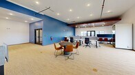Located at 303 Airway Blvd in Belgrade, Montana, this single-floor co-working space offers a thoughtfully designed, professional environment suited for a range of working styles. The open main area is expansive and well-lit, featuring a mix of dedicated desks with privacy screens, round collaborative tables paired with ergonomic leather chairs, and a comfortable lounge seating arrangement — all set on neutral-toned carpeting that runs throughout the space. A striking bold-blue accent wall anchors one side of the main area, contrasting beautifully with crisp white walls and rich wood-slatted ceiling panels that add warmth and architectural character. Pendant and recessed lighting, along with exposed ceiling elements and track lighting, contribute to a dynamic yet focused ambiance. Large windows along the perimeter flood the interior with natural light and offer open views of the surrounding Montana landscape. A private phone booth provides a dedicated space for focused calls or video meetings. Additional enclosed rooms include a clean, minimalist private office and a dedicated conference room featuring a large wooden table, leather rolling chairs, and roller shade windows. The overall design balances collaborative energy with the privacy needs of modern professionals, making this a versatile and well-appointed co-working destination.