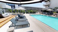This rooftop amenity deck delivers a polished, resort-inspired outdoor experience elevated above the surrounding cityscape. The centerpiece is a generously sized rectangular pool with crystal-clear turquoise water, framed by a wide, light-toned concrete surround that provides ample space for relaxing and socializing. A row of sleek, modern sun loungers in a cool grey finish lines the poolside, accompanied by a bold black-and-white striped umbrella that adds a graphic, contemporary touch. Potted columnar trees line the perimeter, introducing a clean, manicured landscaping element that provides both visual interest and a sense of enclosure.

Away from the pool, a warm hardwood deck area offers a more intimate setting, featuring elegantly designed woven cabana daybed loungers. These oversized pieces are outfitted with plush white cushions and patterned accent pillows, creating a comfortable, lounge-like atmosphere ideal for relaxing in the shade. The arched canopy framework of each cabana adds architectural character while providing shelter from the sun. Together, the pool zone and lounge deck create a cohesive, upscale outdoor retreat with an open-air ambiance and sweeping urban views.