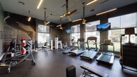 This well-appointed fitness center in Humble, TX offers a comprehensive workout environment designed with both functionality and modern aesthetics in mind. The space features dark-painted walls contrasted by large, floor-to-ceiling windows that line the perimeter, flooding the interior with natural light and providing views of the surrounding greenery. Gold-accented pendant ceiling fans suspended from above add a stylish touch while ensuring air circulation throughout the facility.

The cardio zone is prominently equipped with multiple treadmills, elliptical trainers, a stair climber, and stationary bikes, all positioned to face the expansive windows. Wall-mounted flat-screen televisions are strategically placed for entertainment during workouts. Linear LED lighting fixtures run across the ceiling, providing consistent, focused illumination.

A dedicated strength training area features a variety of cable machines and resistance equipment, complemented by a full-length mirror wall that enhances the sense of space and allows users to monitor their form. Motivational signage is displayed throughout the room, reinforcing an energetic atmosphere. The durable rubber flooring provides traction and safety across all workout zones. Double glass doors provide a welcoming entry point into the facility.