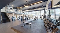 The Athletic Club at The Manor, located at 100 Shepherds Lane in Totowa, NJ, is a expansive two-floor fitness facility with equal floor space distributed across both levels, connected by a sleek staircase with glass railings. The design blends industrial and modern aesthetics, featuring exposed brick walls, dark steel beams, and open ceilings with visible ductwork accented by contemporary circular and linear pendant lighting. Large floor-to-ceiling windows flood both floors with natural light while offering views of the surrounding neighborhood.

The ground floor houses a comprehensive strength training area equipped with a wide selection of selectorized machines, free weights, weight racks, and benches, all set on durable rubber flooring. A dedicated functional training zone features artificial turf, battle ropes, a punching bag, sleds, and barbells. Mirrored walls expand the sense of space throughout, and wall-mounted flat-screen TVs provide entertainment.

The upper level features a cycling studio with stationary bikes positioned alongside panoramic windows, as well as additional cardio equipment and strength machines. A glass-railed mezzanine overlooks the ground floor, reinforcing the facility's open, interconnected feel. A lounge area with indoor plants adds a welcoming touch. The overall atmosphere is modern, energetic, and well-equipped for a full range of fitness activities.
