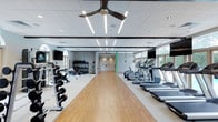 This fitness center offers a spacious, single-floor layout designed to accommodate a wide range of workout needs. The space features a clean, modern aesthetic with a drop ceiling fitted with linear LED lighting and ceiling fans that ensure consistent illumination and air circulation throughout. The flooring transitions between durable wood-toned vinyl and cushioned surfaces, providing appropriate support for both cardio and strength training activities. Along one side of the room, a row of treadmills and elliptical machines is neatly arranged, each positioned to face large windows that allow ample natural light to fill the space and offer views of the surrounding greenery. On the opposite side, a comprehensive free weights section includes barbells, dumbbells, kettlebells, and medicine balls organized on tiered racks, supplemented by wall-mounted mirrors to assist with form and technique. Suspension training equipment is also available, adding functional fitness options. Wall-mounted flat-screen televisions are positioned for visibility throughout the cardio zone. A connecting corridor leads to additional areas, suggesting further amenities beyond the main gym floor. The overall ambiance is bright, open, and well-organized, creating an inviting environment suited for a broad range of fitness activities.