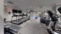 This fitness center is designed to accommodate a comprehensive range of workout routines within a clean, neutral-toned environment. The space is finished with durable speckled rubber flooring throughout, offering both safety and practicality for exercise use. Recessed lighting and ceiling fans are installed across the ceiling, ensuring consistent illumination and air circulation during workouts.

The cardio zone features multiple treadmills and elliptical machines, each equipped with individual screens, arranged along one wall beneath wall-mounted flat-screen televisions. Adjacent to this area, stability balls and open floor space allow for stretching and functional training activities.

The strength training section includes a multi-station cable machine, a full rack of dumbbells on tiered stands, and an adjustable workout bench. Large mirrors line the walls of this zone, visually expanding the space and allowing for form monitoring during exercises.

Near the entrance, a rowing machine and additional equipment are positioned on protective mats. A dedicated alcove provides a touchscreen-enabled water fountain and bottle-filling station alongside a louvered door leading to a restroom or utility area. Signage and a wall clock contribute to the facility's organized, functional atmosphere.