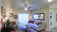 This single-floor residence offers a well-appointed living area designed for both comfort and functionality. The living room serves as the central gathering space, anchored by a neutral-toned sofa dressed with colorful accent pillows and paired with a sleek glass-top coffee table. Warm beige walls create a welcoming ambiance, complemented by wood-style flooring that runs throughout the space.

A large framed mirror mounted on the wall adds visual depth, while a stylish floor lamp provides warm supplemental lighting. The ceiling features a multi-blade ceiling fan with an integrated light fixture, ensuring both air circulation and overhead illumination. A flush-mount ceiling light further brightens the room.

Large sliding glass doors lined with vertical blinds allow an abundance of natural light to fill the space and appear to open onto an outdoor balcony or patio, offering views to the exterior. A hallway visible from the living area leads to an adjacent bedroom, suggesting a logical and efficient floor plan. The overall aesthetic is clean, modern, and cohesive, with neutral tones and practical fixtures throughout.