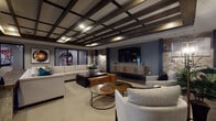 This single-floor amenity space is thoughtfully designed to serve as a premier entertainment and social hub. The layout is divided into distinct yet open zones, each serving a specific leisure function while contributing to a cohesive, upscale aesthetic.

The lounge area is anchored by a striking coffered ceiling with dark wood beams and recessed lighting, adding architectural depth and warmth overhead. A large sectional sofa, accent chairs, and layered coffee tables surround a wall-mounted television set against a blue feature wall with stacked stone accents, creating an inviting media and relaxation zone. Bold artwork adds a curated, contemporary touch throughout.

Adjacent to the lounge, the games area features a substantial rustic wood billiards table suspended beneath an industrial-style pendant light. A dedicated card or game table with surrounding seating offers additional entertainment options. The flooring throughout this zone consists of large-format slate-toned tiles that ground the space with an earthy, durable finish.

A bar-height counter with seating provides a casual gathering point, while stone-clad columns punctuate the space, reinforcing the natural material palette. Windows and glass-panel walls allow views between zones and bring in ambient light. An exterior-facing entrance with glass doors connects the space to adjacent areas, completing this well-appointed communal retreat.