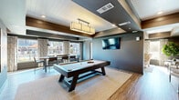 Located at 3262 S Railroad St in Phenix City, Alabama, this single-floor community clubhouse offers a thoughtfully designed gathering space that blends modern aesthetics with welcoming comfort. The interior is finished with warm wood-look flooring, deep gray walls, and coffered ceilings with white shiplap accents, creating a sophisticated yet inviting atmosphere throughout.

The main game and lounge area serves as the centerpiece of the space, featuring a full-size pool table illuminated by a statement pendant fixture, wall-mounted flat-screen TV, and ample open floor space. Adjacent to this is a relaxed lounge nook with upholstered seating, decorative wall art, and accent furnishings that encourage casual conversation.

A cozy sitting room bathed in natural light from large windows offers wingback chairs, a camel-toned sofa, and curated landscape artwork, providing a quieter retreat within the space. The lobby entry area showcases eclectic décor including a circular mirror, driftwood console, and decorative wall-mounted pieces that establish a distinctive character.

A communal dining zone features a long wooden table surrounded by industrial-style chairs, framed by large windows overlooking an outdoor pool area. A banquette lounge lined with shiplap walls and decorative antler mounts rounds out the space, accompanied by a refreshment counter. Natural light and outdoor views are prominent throughout.