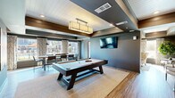 Located at 3262 S Railroad St in Phenix City, Alabama, this single-floor community clubhouse offers a thoughtfully designed gathering space that blends modern aesthetics with welcoming comfort. The interior is finished with warm wood-look flooring, deep gray walls, and coffered ceilings with white shiplap accents, creating a sophisticated yet inviting atmosphere throughout.

The main game and lounge area serves as the centerpiece of the space, featuring a full-size pool table illuminated by a statement pendant fixture, wall-mounted flat-screen TV, and ample open floor space. Adjacent to this is a relaxed lounge nook with upholstered seating, decorative wall art, and accent furnishings that encourage casual conversation.

A cozy sitting room bathed in natural light from large windows offers wingback chairs, a camel-toned sofa, and curated landscape artwork, providing a quieter retreat within the space. The lobby entry area showcases eclectic décor including a circular mirror, driftwood console, and decorative wall-mounted pieces that establish a distinctive character.

A communal dining zone features a long wooden table surrounded by industrial-style chairs, framed by large windows overlooking an outdoor pool area. A banquette lounge lined with shiplap walls and decorative antler mounts rounds out the space, accompanied by a refreshment counter. Natural light and outdoor views are prominent throughout.