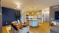This thoughtfully designed single-floor residence offers a seamless open-concept layout that connects the kitchen, dining, and living areas into one cohesive, welcoming space. The kitchen is a standout feature, outfitted with light wood cabinetry, stainless steel appliances, a spacious center island with granite-style countertops, and pendant lighting that adds a warm, modern touch. A bold navy accent wall anchors the dining area, where warm hardwood-style flooring flows throughout the main living spaces. The living room is bright and inviting, featuring a sliding glass door that opens to an outdoor balcony, allowing natural light to fill the room. Two bathrooms serve the residence — the primary bath includes a wide granite-topped double vanity and walk-in closet access, while the second bath features a full tub-shower combo with clean, contemporary finishes. The bedroom is a calm retreat with plush carpeting, a ceiling fan, natural light from a large window, and a teal accent wall that adds depth and character. Large-scale artwork displayed throughout the dining and living areas contributes to an elevated, curated aesthetic that runs consistently across the entire space.