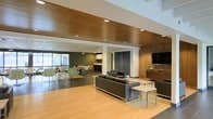 This single-floor commercial space offers a thoughtfully designed open-concept layout that blends functionality with contemporary style. The lounge area serves as a focal point, furnished with sleek dark upholstered sofas and accent chairs arranged around low coffee tables, anchored by light hardwood flooring that adds warmth to the setting. A wall-mounted flat-screen display and built-in wood cabinetry provide both practicality and a polished finish to this area.

Recessed lighting is embedded throughout the striking wood-paneled ceiling, casting a warm, even glow across the space. Transitioning from the lounge, a casual dining or breakroom area features round tables paired with modern molded chairs in neutral tones, positioned near large windows that allow natural light to filter in, contributing to an open and airy atmosphere.

The space also includes a compact kitchenette or service counter tucked neatly along one wall, and dark accent walls provide contrast against the lighter flooring and ceiling elements. A corridor with institutional gray doors leads further into the facility, suggesting additional private or functional rooms beyond the main open area. Overall, the space conveys a professional yet welcoming ambiance well-suited for a corporate lounge, hospitality, or community gathering environment.