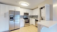This single-floor residence presents a clean, modern aesthetic characterized by light-toned tile flooring laid throughout in a wood-plank style, neutral warm-gray walls, and crisp white baseboards. The open-concept living and dining area is generously proportioned, offering a seamless flow toward the kitchen and access to a sliding glass door that invites natural light and views of the surrounding greenery.

The kitchen is well-appointed with white shaker-style cabinetry, a mosaic tile backsplash, granite-look countertops, and a full suite of stainless steel appliances including a refrigerator with ice dispenser, range, microwave, and dishwasher. A dedicated in-unit stacked washer and dryer is conveniently tucked behind louvered doors within the kitchen area.

The bedrooms are comfortably sized, each featuring natural light from windows with blinds. The primary bedroom connects to a walk-in closet with built-in shelving and an en-suite bathroom featuring a tub-shower combination with floor-to-ceiling tile surround, a dark wood vanity, and a granite countertop. A hallway provides access to additional rooms and storage, contributing to the unit's practical and well-organized layout.