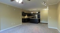 This single-floor residence offers an open-concept design that seamlessly connects the living area and kitchen, creating a cohesive and functional layout. The kitchen is a standout feature, showcasing rich dark cabinetry that contrasts elegantly with granite countertops. A central island provides additional workspace and casual seating, making it an ideal hub for both cooking and entertaining. Stainless steel appliances, including a refrigerator, range, and microwave, complete the modern kitchen setup. Track lighting and pendant lights illuminate the kitchen, while a ceiling fan in the living area ensures comfortable airflow throughout. Crown molding adds a refined architectural touch along the ceilings, lending the space a polished, finished look. Plush carpeting in the living area creates a warm and inviting atmosphere underfoot. The open floor plan allows for flexible furniture arrangements, giving the space a sense of openness and adaptability. Neutral wall tones provide a versatile backdrop that complements the bold cabinetry and dark finishes throughout, making this a well-appointed and thoughtfully designed living space.