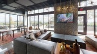 This expansive single-floor amenity space is designed with a contemporary industrial aesthetic, combining polished concrete floors, exposed black ceilings with open ductwork, and warm wood accents throughout. The layout seamlessly connects multiple distinct activity zones while maintaining an open, cohesive feel.

The lounge area anchors the space with deep sectional seating positioned around a stone accent wall mounted with a large flat-screen TV. Floor-to-ceiling glass sliding doors flood the room with natural light and provide direct access to an outdoor terrace, creating a fluid indoor-outdoor connection.

The game room features a billiards table, ping-pong table, shuffleboard, and a Jenga station, all set against light wood-paneled walls with curated artwork. Large sliding barn-style doors allow the space to be divided or opened as needed.

A stylish bar and café area showcases reclaimed wood cabinetry, Edison bulb pendant lighting with a colorful overhead installation, bar seating, and built-in appliances — ideal for entertaining or casual gatherings.

Throughout the space, modern geometric light fixtures, eclectic artwork, and natural materials including stone, wood, and woven textiles contribute to a vibrant yet welcoming atmosphere.