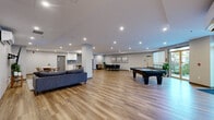 This generously sized single-floor amenity space offers an open-concept layout designed for both relaxation and social interaction. The main living area is anchored by a large sectional sofa and a wall-mounted flat-screen TV, complemented by a ductless mini-split HVAC unit for year-round comfort. Recessed lighting throughout provides a clean, modern ambiance, while warm-toned hardwood-style flooring runs seamlessly across the entire space, lending a cohesive and inviting feel.

A full kitchen is positioned toward the rear, featuring white upper cabinetry, a grey subway tile backsplash, and stainless steel appliances including a full-sized refrigerator. Adjacent to the kitchen, a casual dining table with bar-height seating offers a practical gathering spot. A separate dining area accommodates a long table with multiple chairs, suitable for group meals or meetings.

The space also includes a dedicated recreation zone with a full-sized pool table, adding an entertainment dimension to the layout. A secondary seating area near a large window allows natural light to filter in, creating a bright and welcoming corner. A hallway with double doors provides access to the rest of the building. The overall design is modern and functional, making this a versatile communal space well-suited for a variety of uses.
