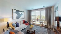 This single-floor residence offers a thoughtfully designed layout that balances comfort and modern style across its key living spaces. The open-concept living and dining area serves as the heart of the home, featuring warm hardwood-style flooring, large windows with horizontal blinds and floor-length drape panels, and generous natural light with outdoor views. Bold abstract artwork accents the neutral walls, while a plush area rug and round coffee table anchor the sitting area. The well-equipped kitchen is outfitted with stainless steel appliances including a refrigerator, range, microwave, dishwasher, and a wine cooler. White shaker-style cabinetry, granite countertops, an undermount sink, and pendant lighting complete the contemporary look. The primary bedroom features carpet flooring, a statement drum chandelier in gold, large windows, and direct access to a spacious en-suite bathroom with a double-sink vanity, granite counters, tile flooring, a walk-in closet, and a glass-enclosed shower with a built-in bench. The second bedroom offers a calming blue accent wall, carpet, and ample natural light. A dedicated in-unit laundry room houses a full-size front-loading washer and dryer with overhead shelving for added storage.