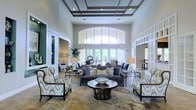 This beautifully appointed single-floor space exudes a refined and welcoming ambiance, designed to impress from the moment you step inside. The centerpiece of the main lounge area is a dramatic arched window wall that floods the interior with abundant natural light, framing lush outdoor greenery beyond. Soaring ceilings with a striking coffered detail overhead add architectural depth and a sense of grandeur to the space.

The seating area is thoughtfully curated, featuring a plush, deep-toned sofa paired with stylishly patterned accent chairs in swirling earth tones, all arranged around a rich dark wood coffee table. A decorative area rug anchors the arrangement, adding warmth and texture to the light tile flooring beneath. Accent lamps and carefully placed greenery contribute to a layered, inviting atmosphere.

Flanking the main area are additional rooms visible through open doorways, hinting at further functional or lounge spaces. The white wainscoting and paneled wall details on either side of the room provide a classic contrast to the contemporary furnishings. The overall design strikes a balance between elegance and comfort, making this space ideal for gathering, relaxing, or socializing in a polished, upscale environment.