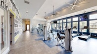 Located at Wolf Ranch Parkway in Georgetown, TX, this fitness center offers a generously sized, single-floor layout designed to support a full range of workout routines. The main gym area is bright and open, featuring soaring ceilings equipped with large industrial-style ceiling fans that promote airflow throughout the space. A full wall of floor-to-ceiling windows along one side floods the room with natural light and provides views of the surrounding outdoor landscape. The main floor houses an extensive array of strength machines, a cable system, a dedicated free weights rack with dumbbells and kettlebells, and multiple cardio machines including treadmills and ellipticals. A bold motivational typography mural covers one wall, energizing the atmosphere with fitness-focused messaging. Large mirrors line sections of the walls, enhancing the sense of space. A separate spin studio features stationary cycling bikes, tall windows with outdoor views, and a striking artistic bicycle mural as a focal point. An additional functional training room provides space for suspension training, a heavy bag, and floor-based exercises. A dedicated entry area includes wall-mounted displays for scheduling and facility information, finished with clean tile flooring and a frosted glass door entry.